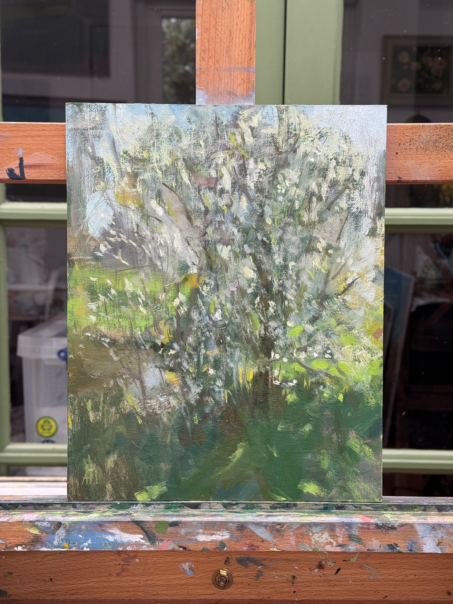 An impressionistic oil painting of a flowering tree in soft greens, whites, and grays, displayed on a wooden easel.