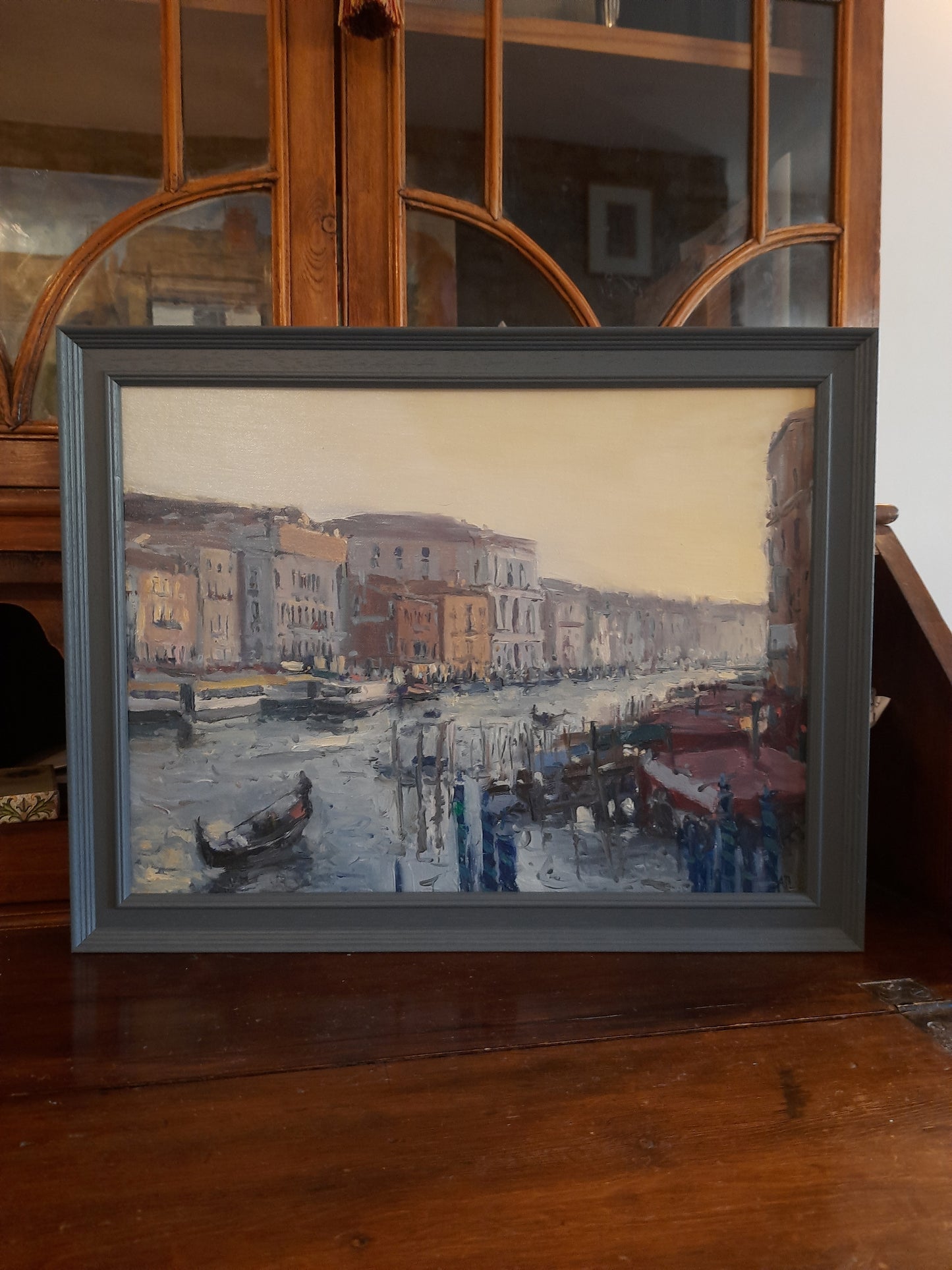 A photograph of the oil painting in a grey frame on a wood secretaire bookcase.