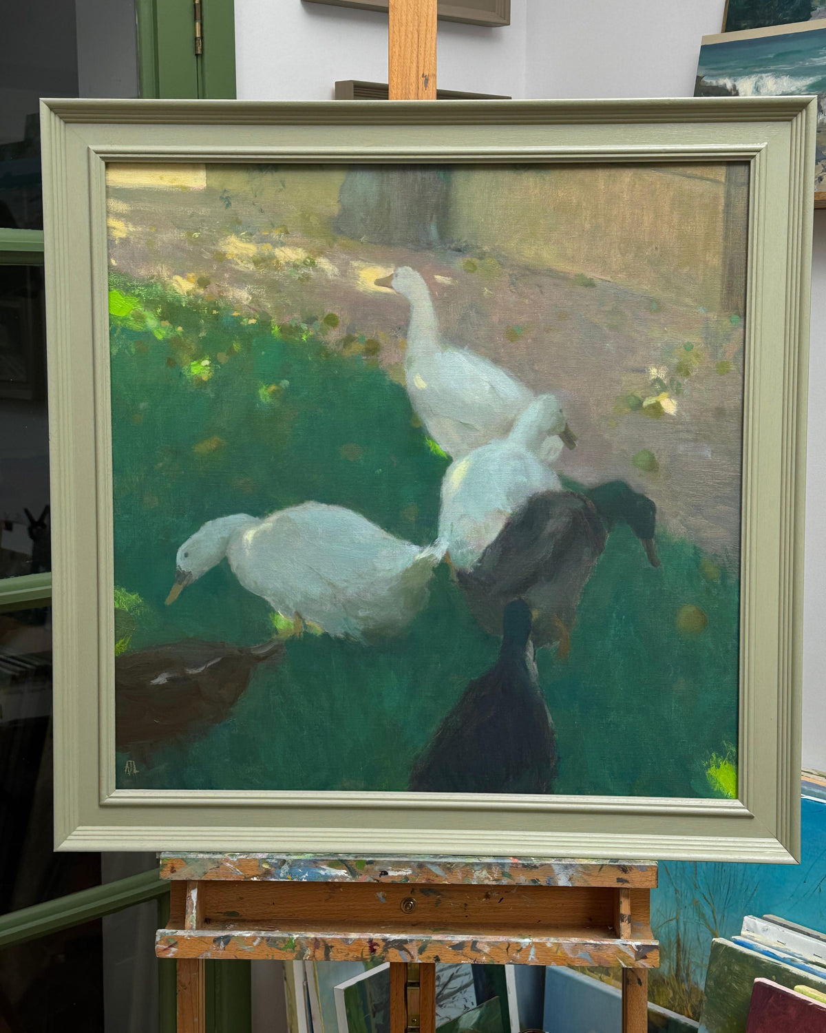 A framed oil painting of several white and dark ducks in a grassy outdoor setting, displayed on a wooden artist easel in a studio.