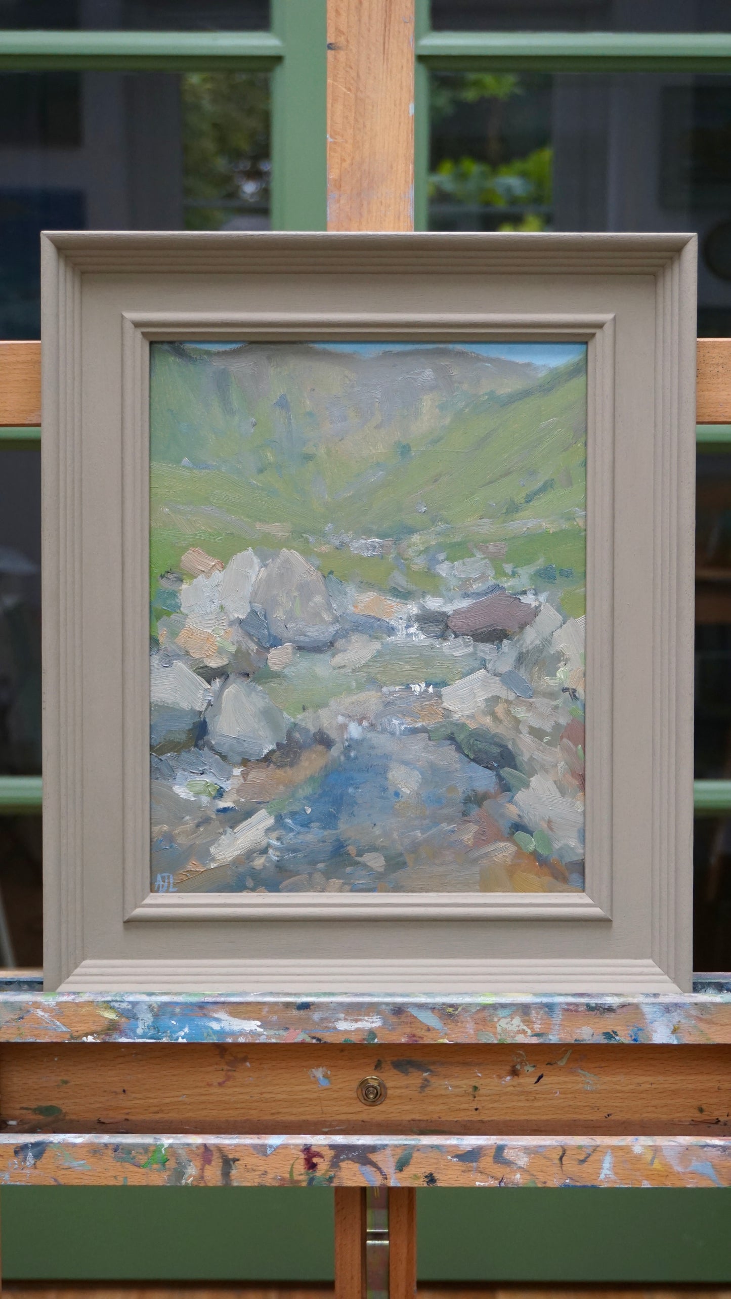 Framed painting of a landscape with mountains and a stream on a wooden surface.