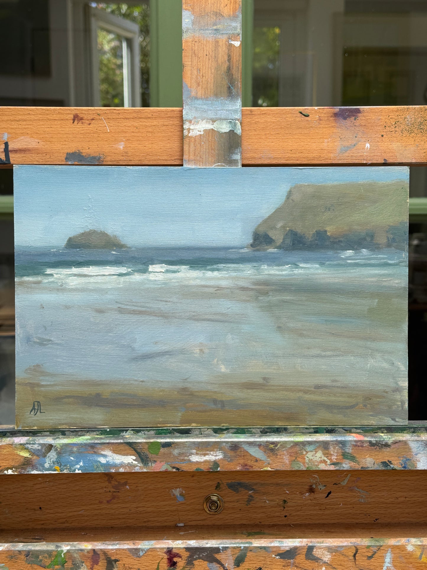 A small landscape painting of a beach and rocky cliffs under a pale sky, displayed on an artist's easel.