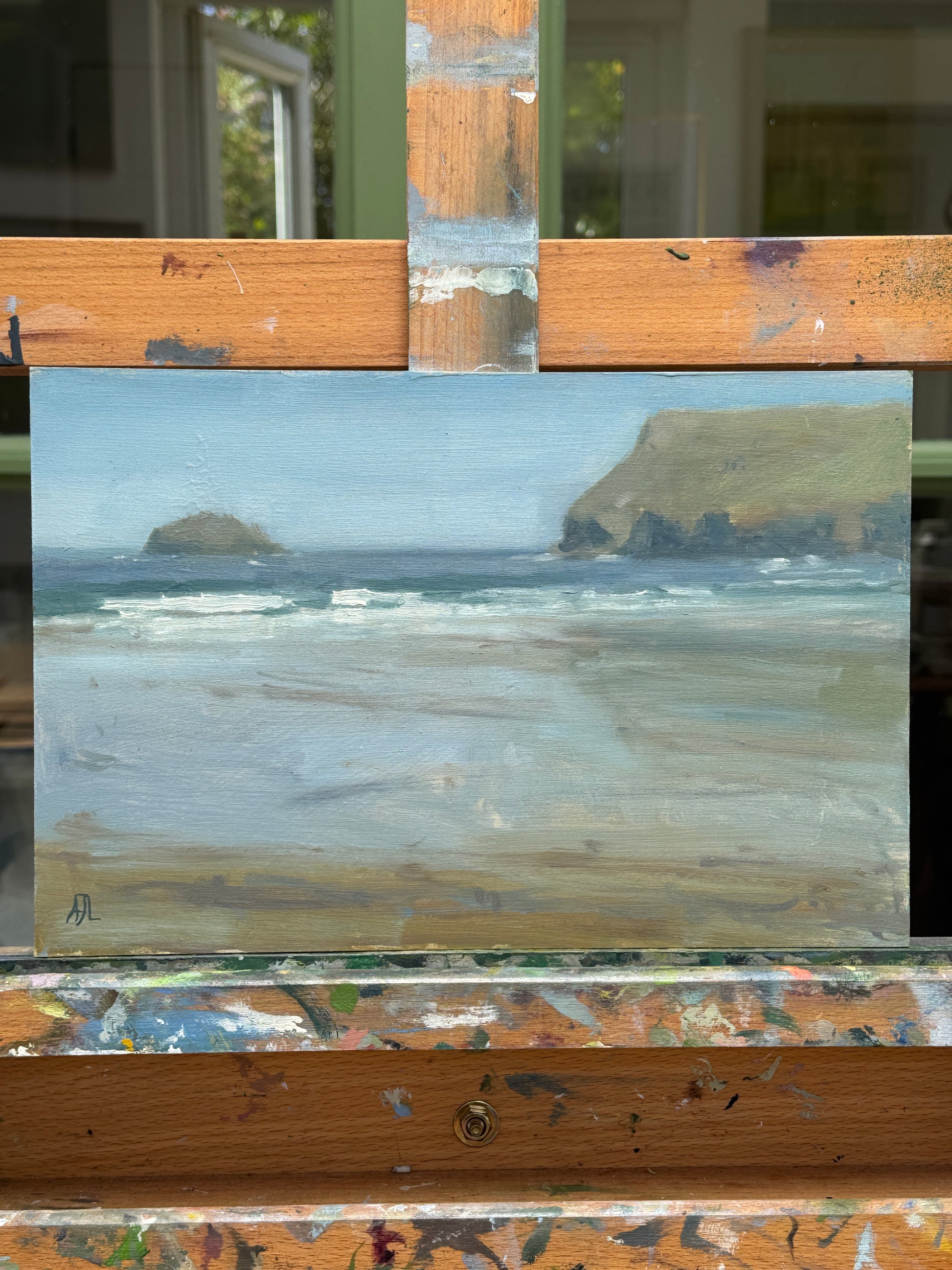 A small landscape painting of a beach and rocky cliffs under a pale sky, displayed on an artist's easel.