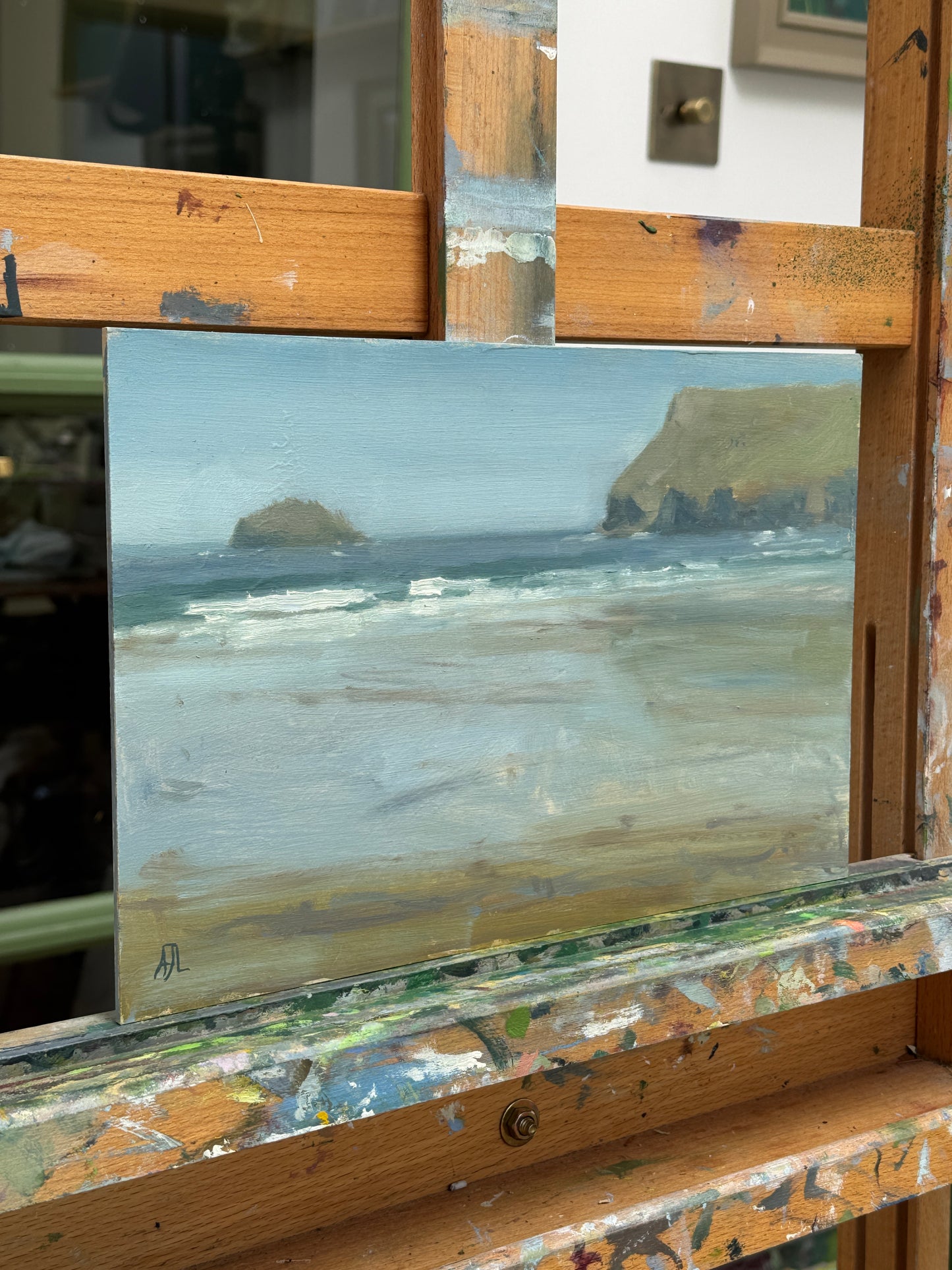 A small landscape oil painting depicting a coastal scene with waves, cliffs, and a rocky island, displayed on a wooden artist easel with paint splatters.