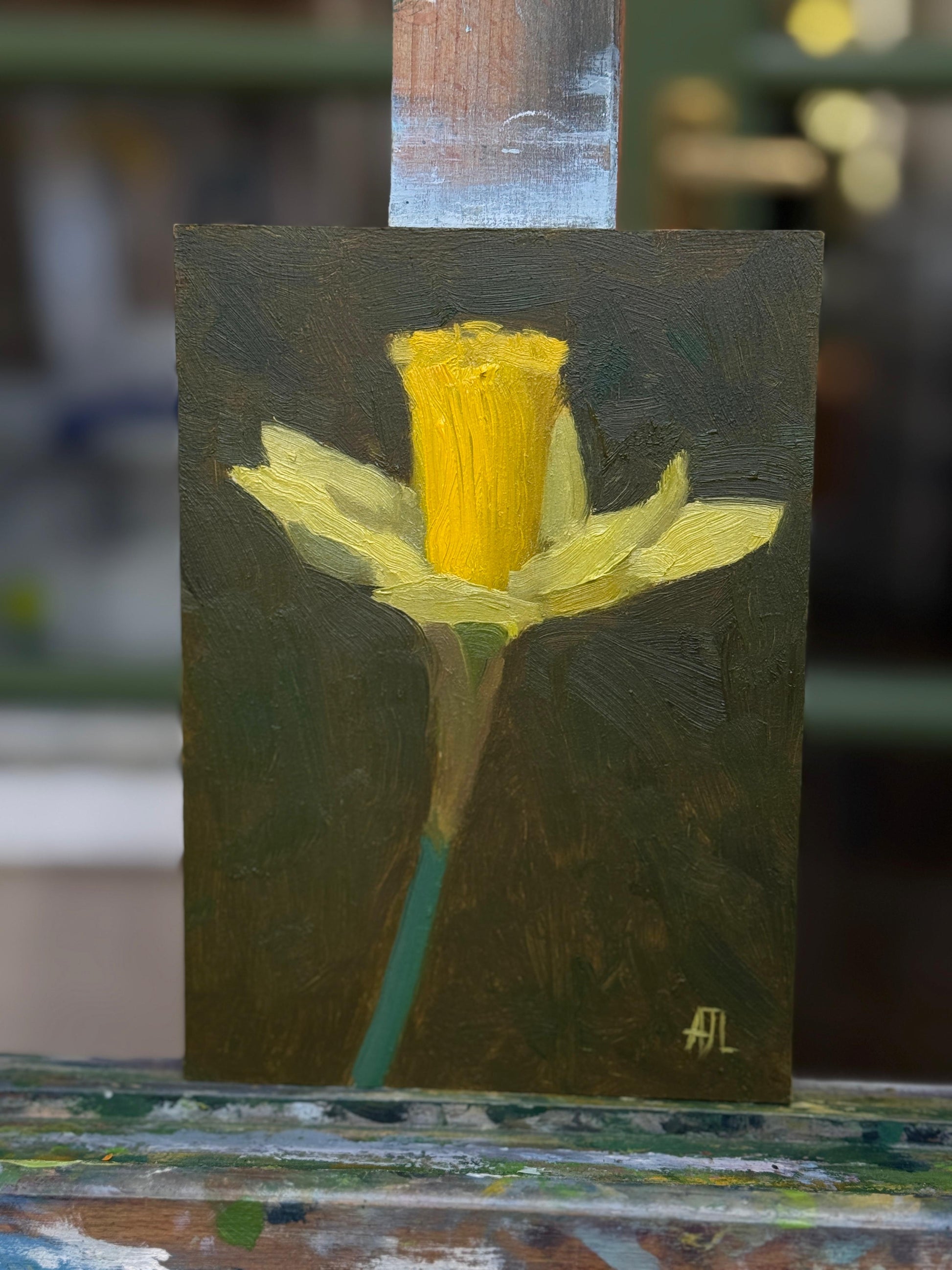 A small vertical oil painting of a yellow daffodil on a dark brown background, displayed on an easel in an artist's studio.