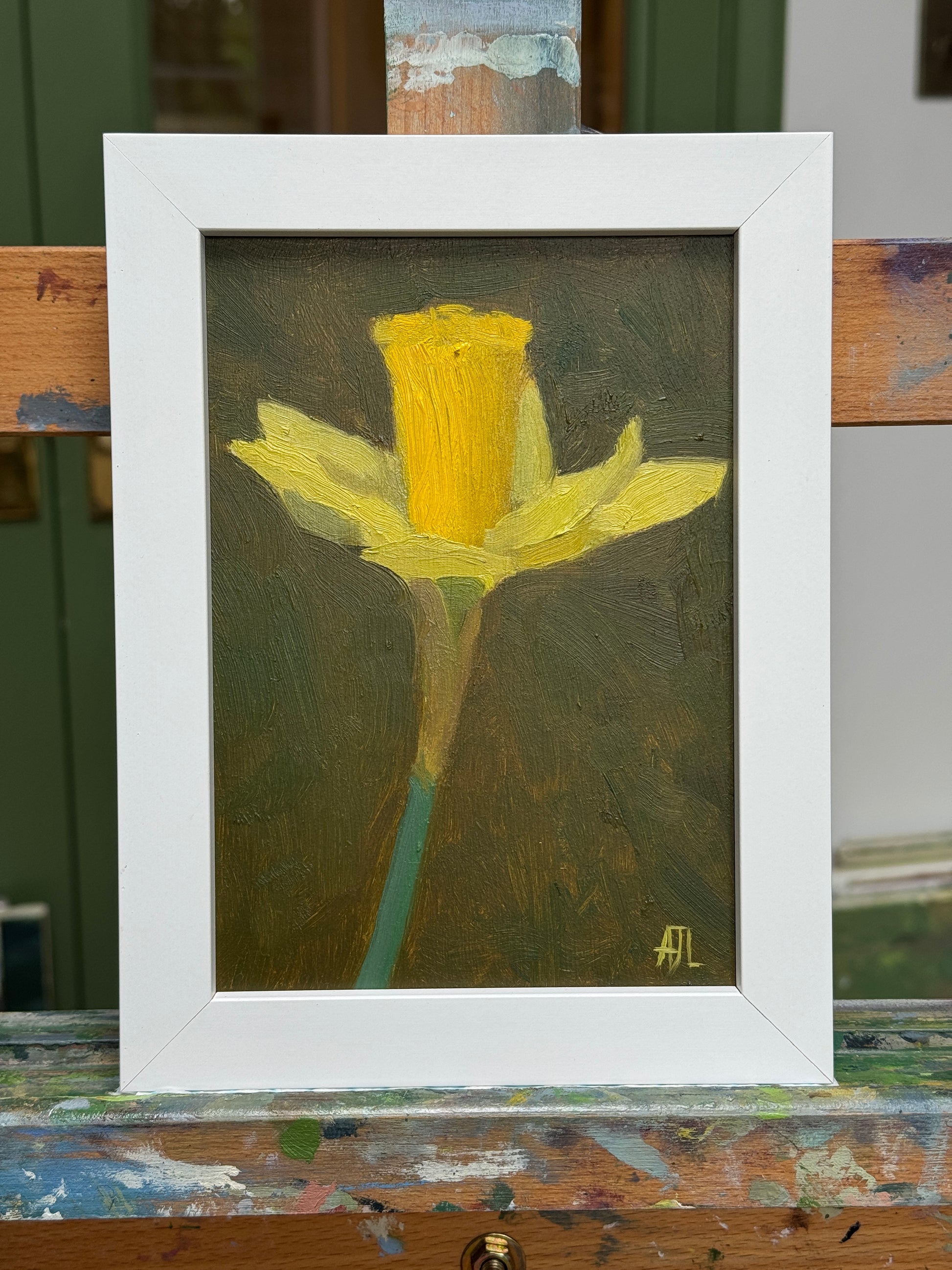 A framed oil painting of a yellow daffodil on a dark textured background, housed in a wide white frame. The frame is shown resting on a wooden artist easel with paint splatters.