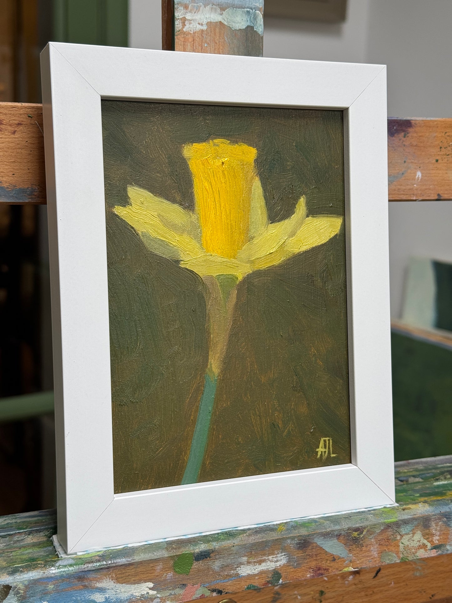 A framed oil painting of a yellow daffodil on a dark olive background, housed in a white frame and resting on a wooden easel.