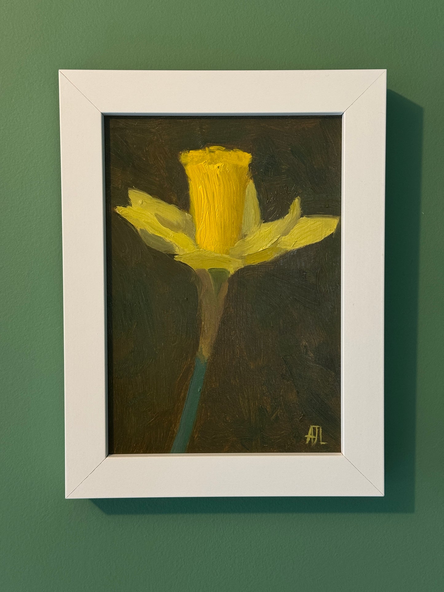 A framed painting of a yellow daffodil on a dark textured background, mounted on a solid green wall.