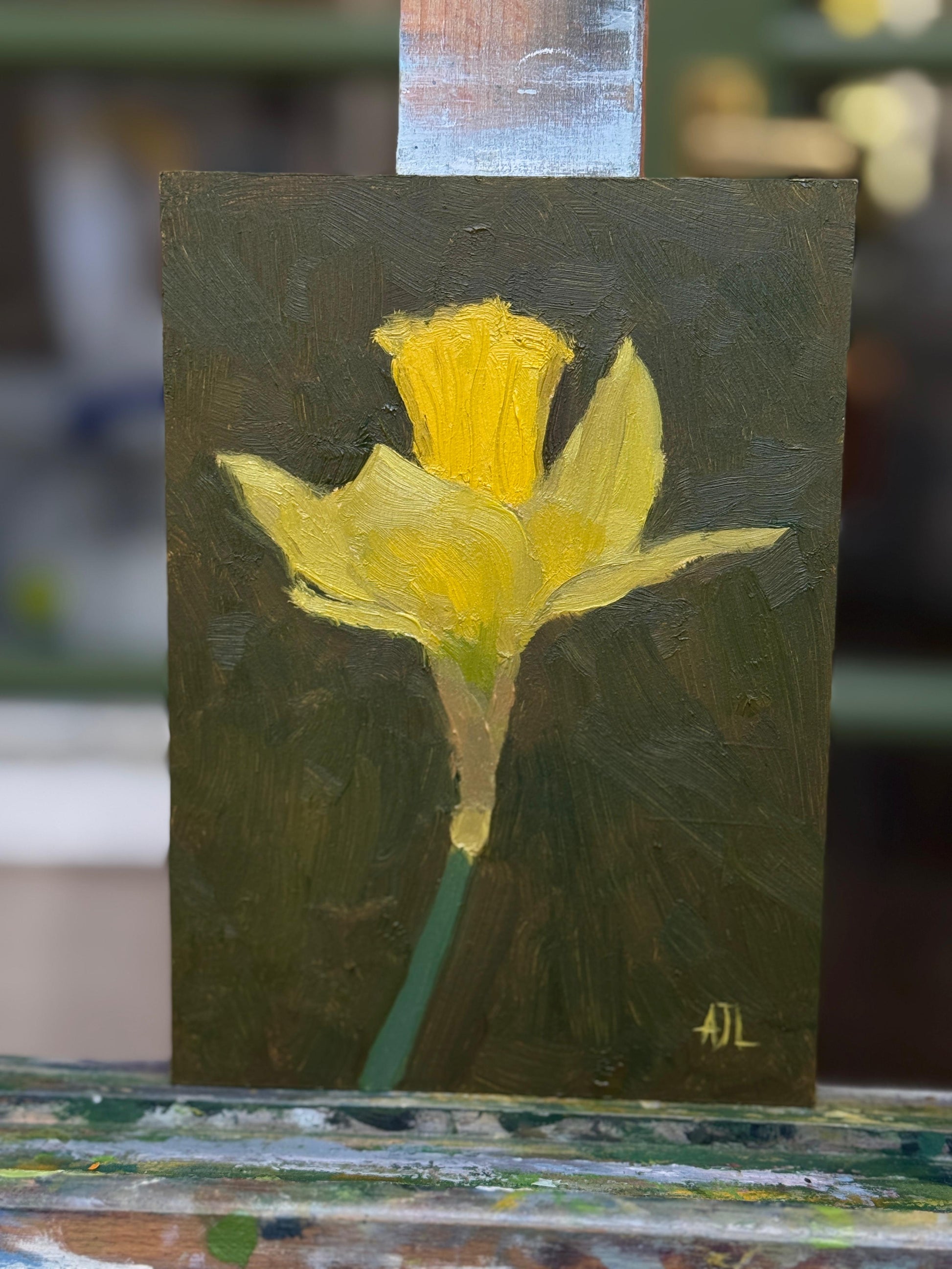 A small painted canvas featuring a bright yellow flower on a dark olive-green background, photographed leaning against a painted wooden surface with a blurred real-world background.