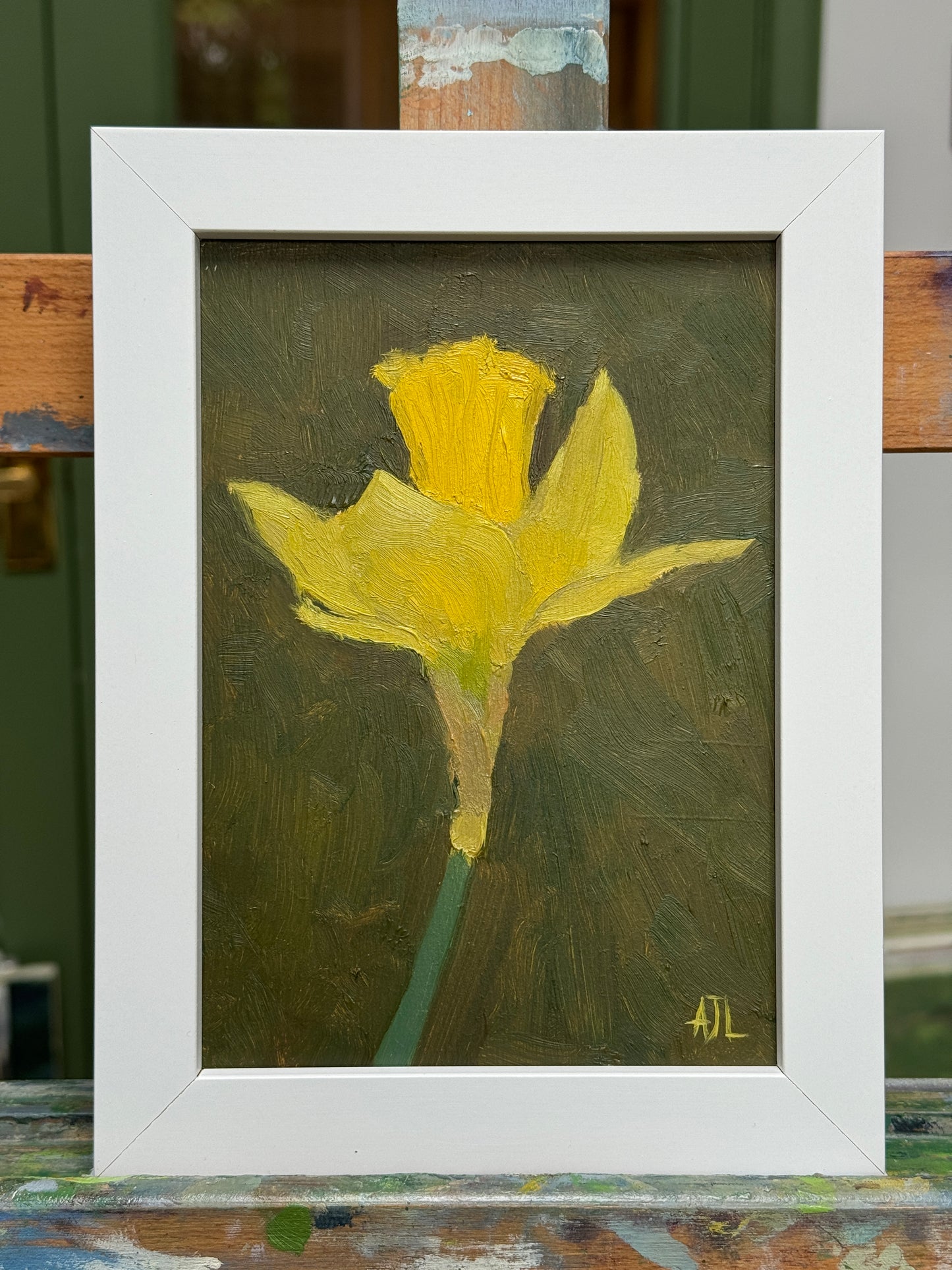 A framed oil painting of a yellow daffodil on a dark olive-green background, housed in a white frame. The painting is displayed resting on a wooden artist easel.