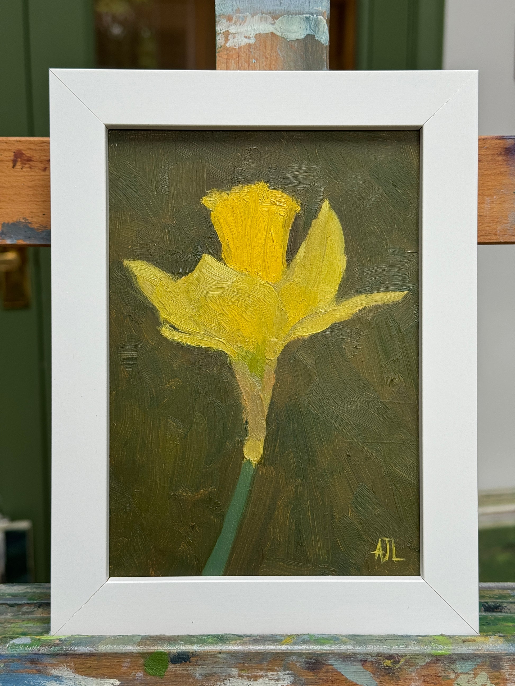 A framed oil painting of a yellow daffodil on a dark olive-green background, housed in a white frame. The painting is displayed resting on a wooden artist easel.