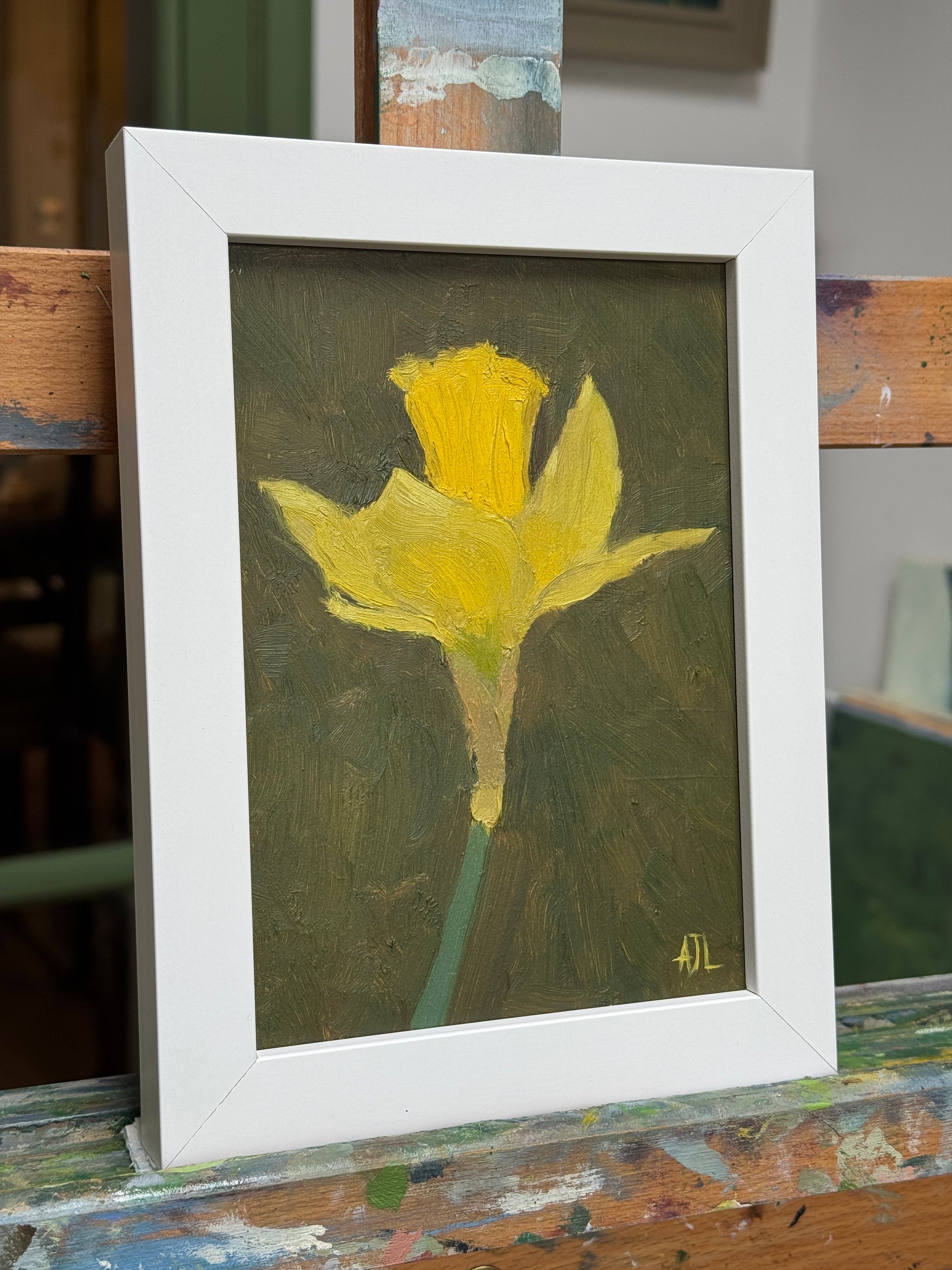 A framed oil painting of a yellow daffodil on a dark textured background, housed in a white frame and displayed on a rustic wooden easel.