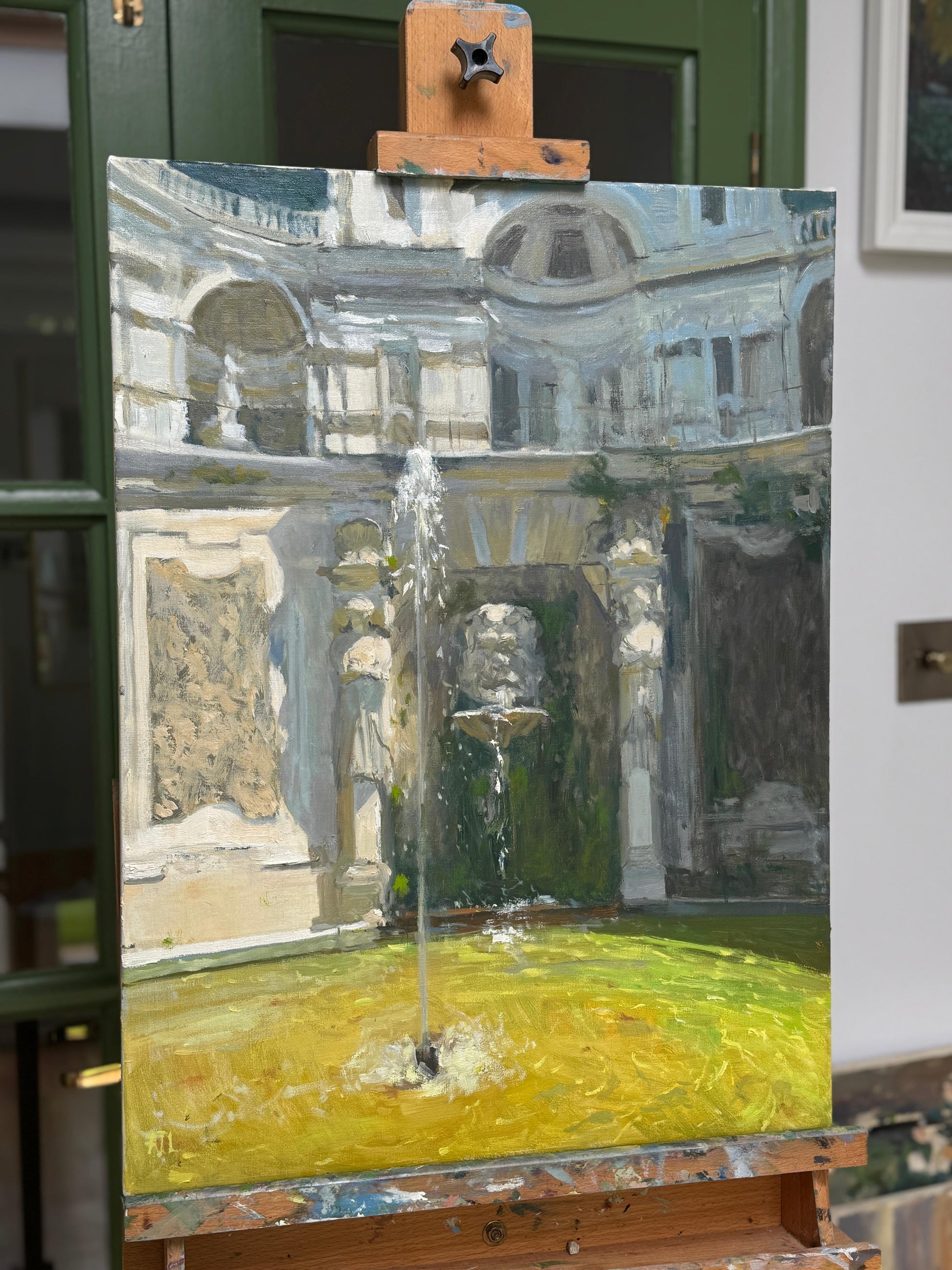 The original oil painting of a courtyard with a fountain on an easel against a green wall.