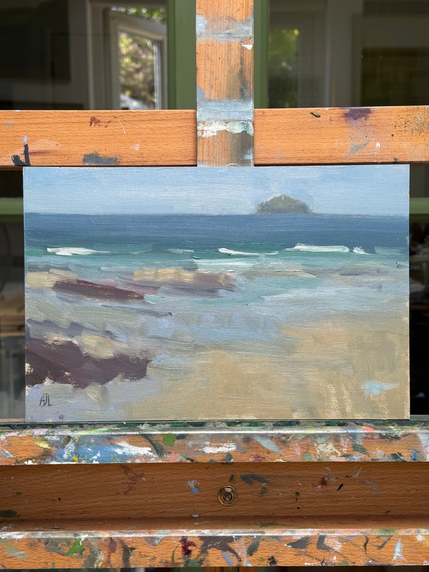 An oil painting depicting a coastal landscape with blue water and sandy beige shore, displayed on a wooden easel with paint splatters.