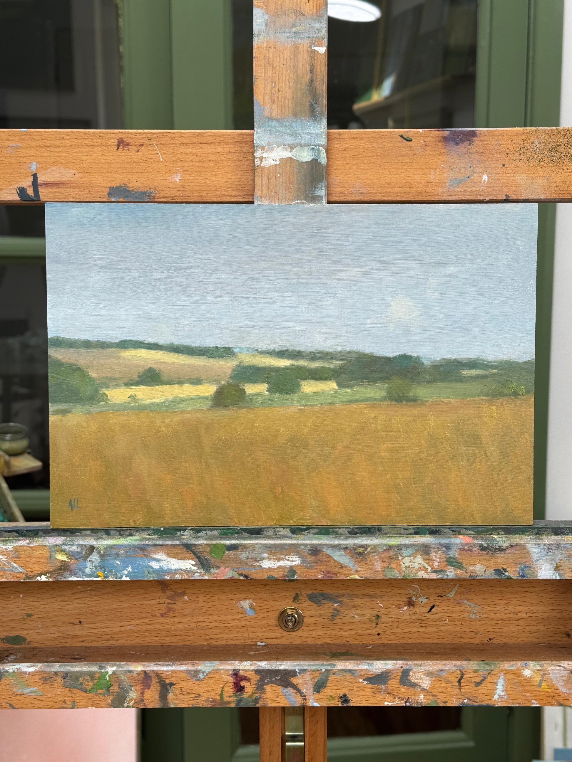 A landscape oil painting mounted on a wooden artist easel. The painting depicts a rolling golden field under a pale blue sky with green hills in the distance.