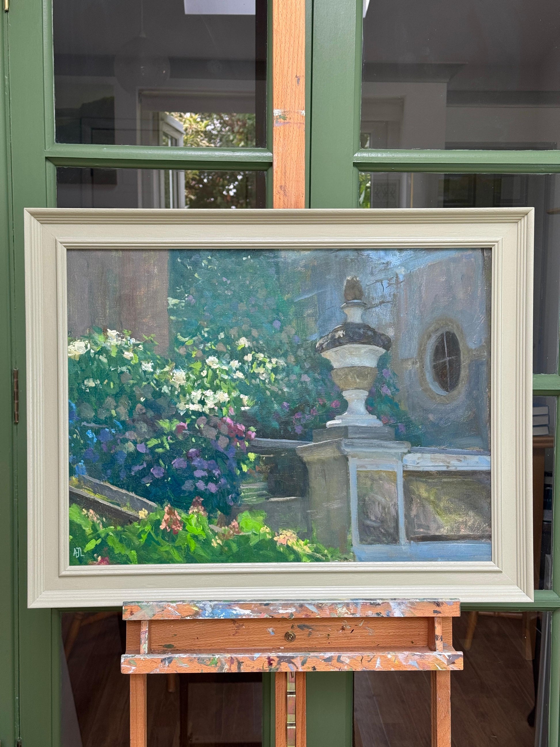 Framed painting of a garden scene on an easel against a window.