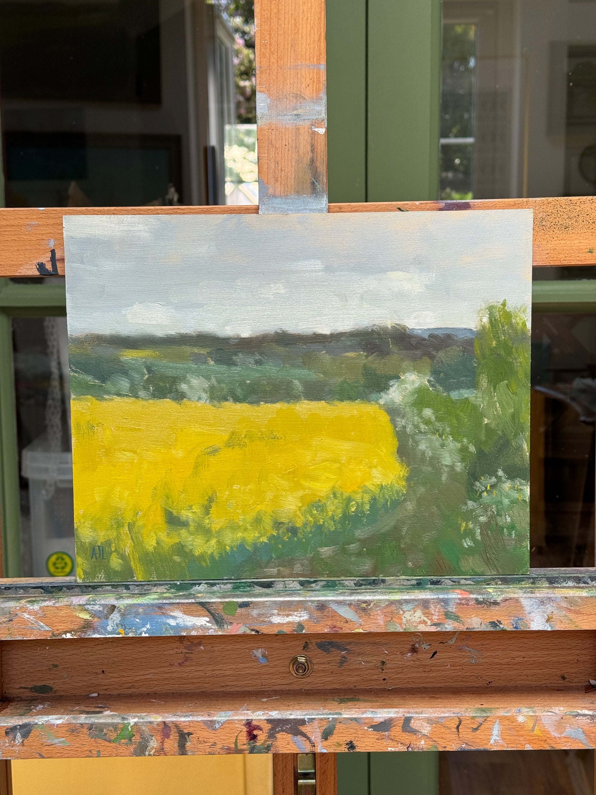 A small impressionistic landscape painting, featuring a bright yellow field under a cloudy gray sky, displayed on a wooden artist easel.