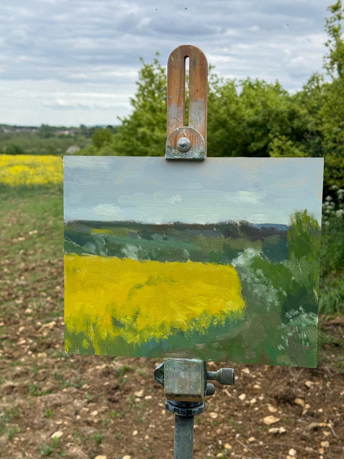 A small painted landscape canvas on an easel stand, showing a yellow field under a cloudy sky, photographed outdoors with trees and ground visible in the background.