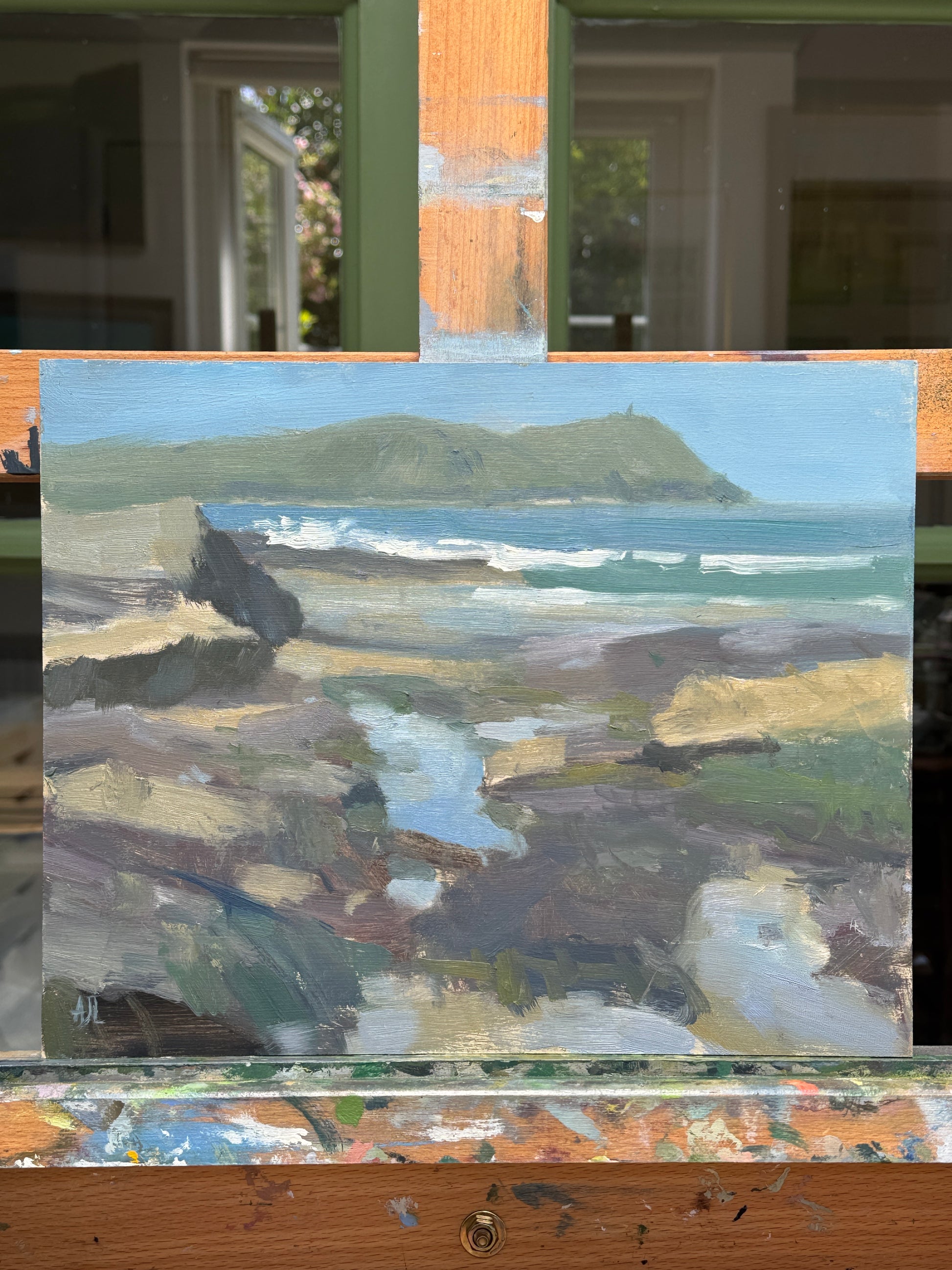 A seascape painting on a canvas, featuring textured brushstrokes of blue, gray, beige, and green, displayed on a wooden easel.