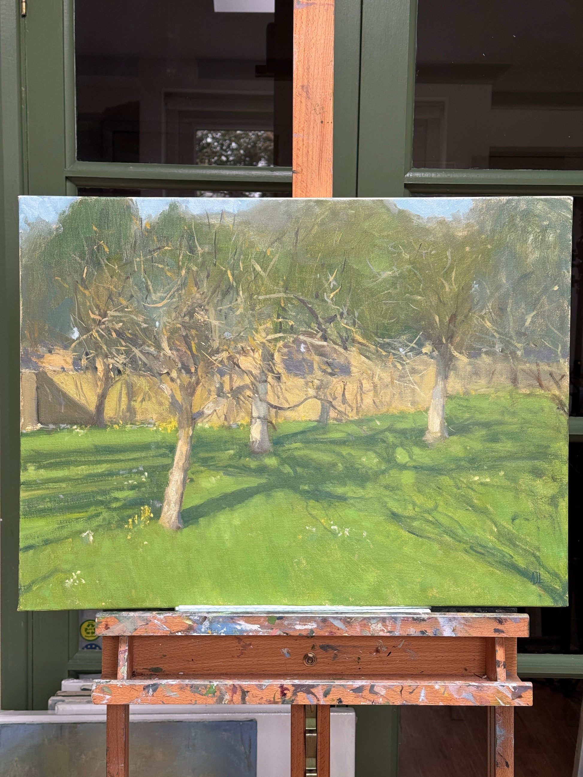 An oil painting of a landscape with green grass, brown tree trunks, and a pale sky, displayed on an easel in an artist's studio.