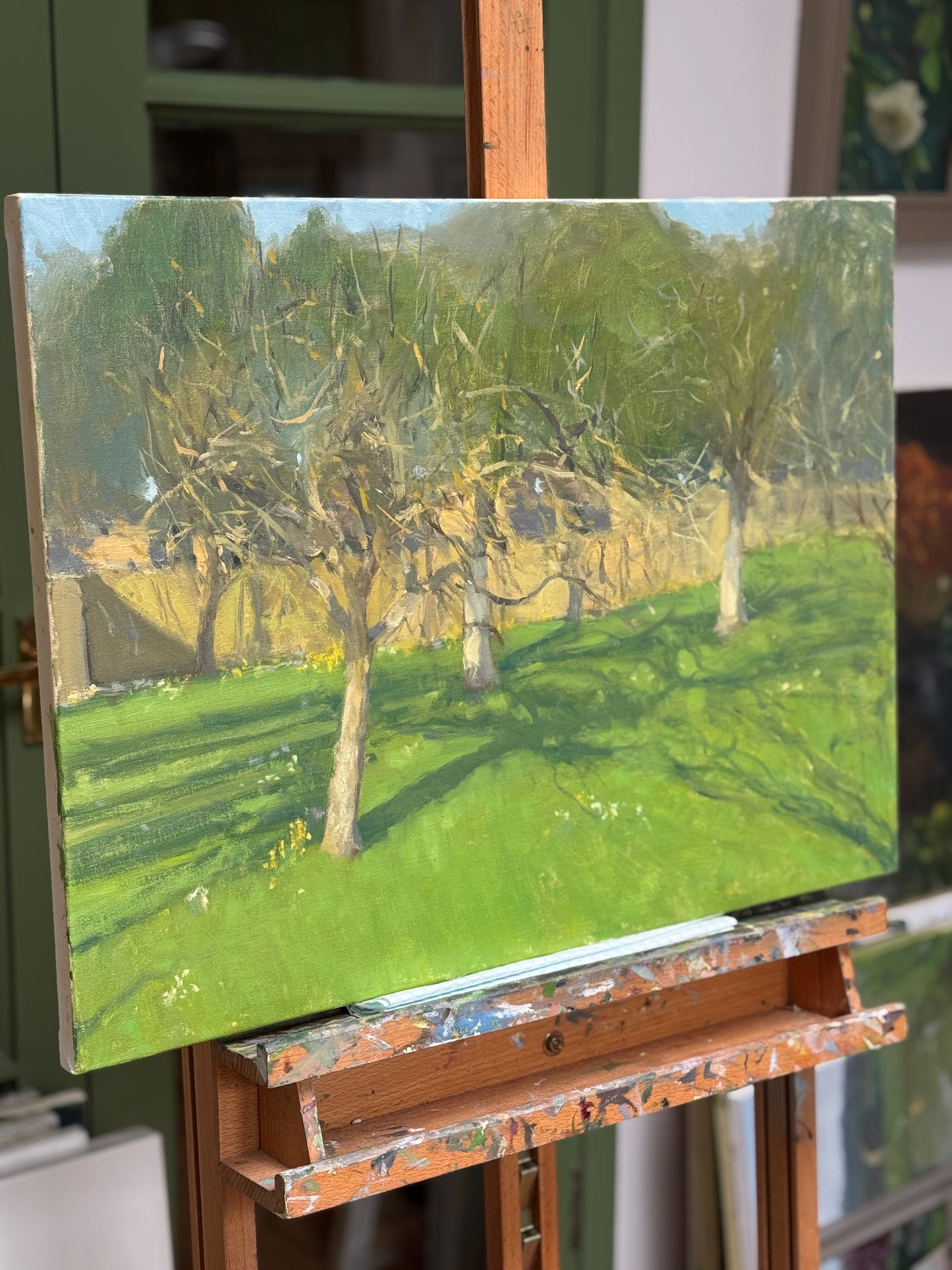 An oil painting of an orchard scene with green grass and trees, displayed on a wooden artist easel in an indoor studio setting.