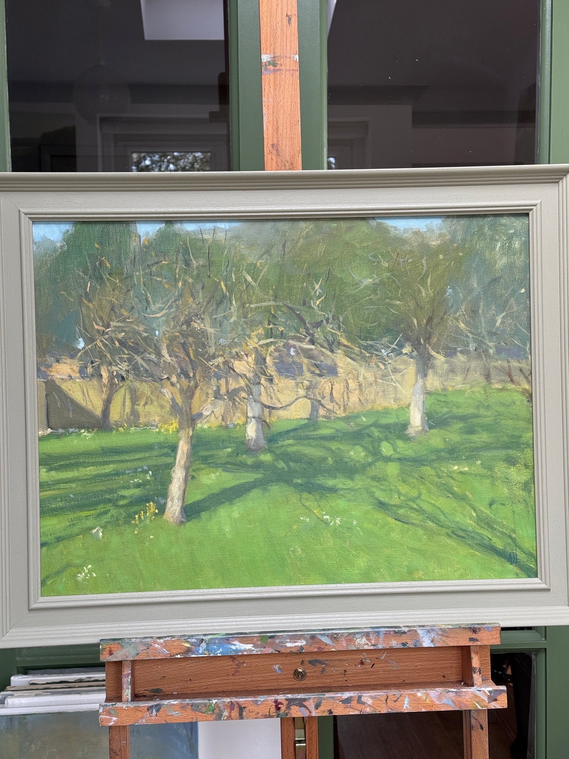 A framed landscape painting featuring a green field with apple trees and a stone wall, displayed in a light sage frame. The painting is photographed in an artist's studio setting on an easel.