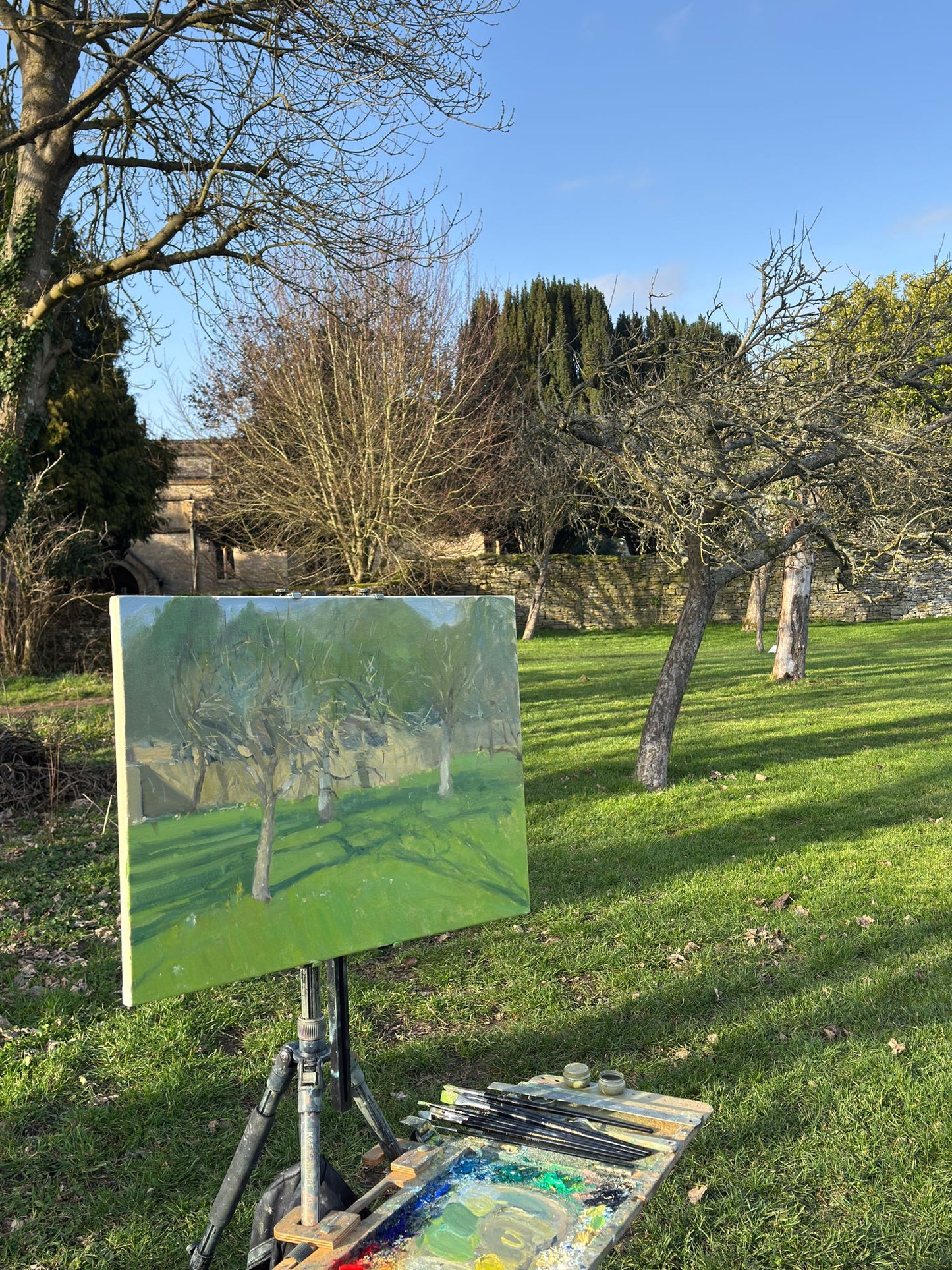 An easel holding a painted canvas of trees in a grassy outdoor landscape, with an artist's palette and brushes on the grass in front. The background shows an orchard with apple trees, grass, and blue sky.