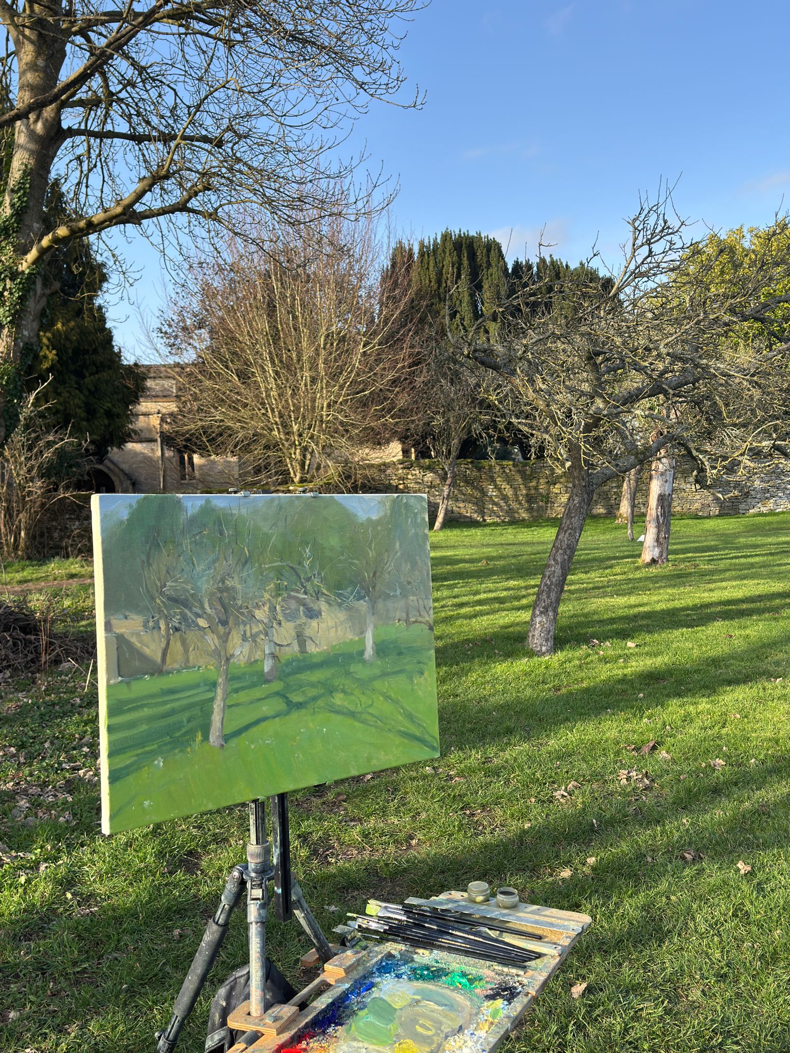 An easel holding a painted canvas of trees in a grassy outdoor landscape, with an artist's palette and brushes on the grass in front. The background shows an orchard with apple trees, grass, and blue sky.
