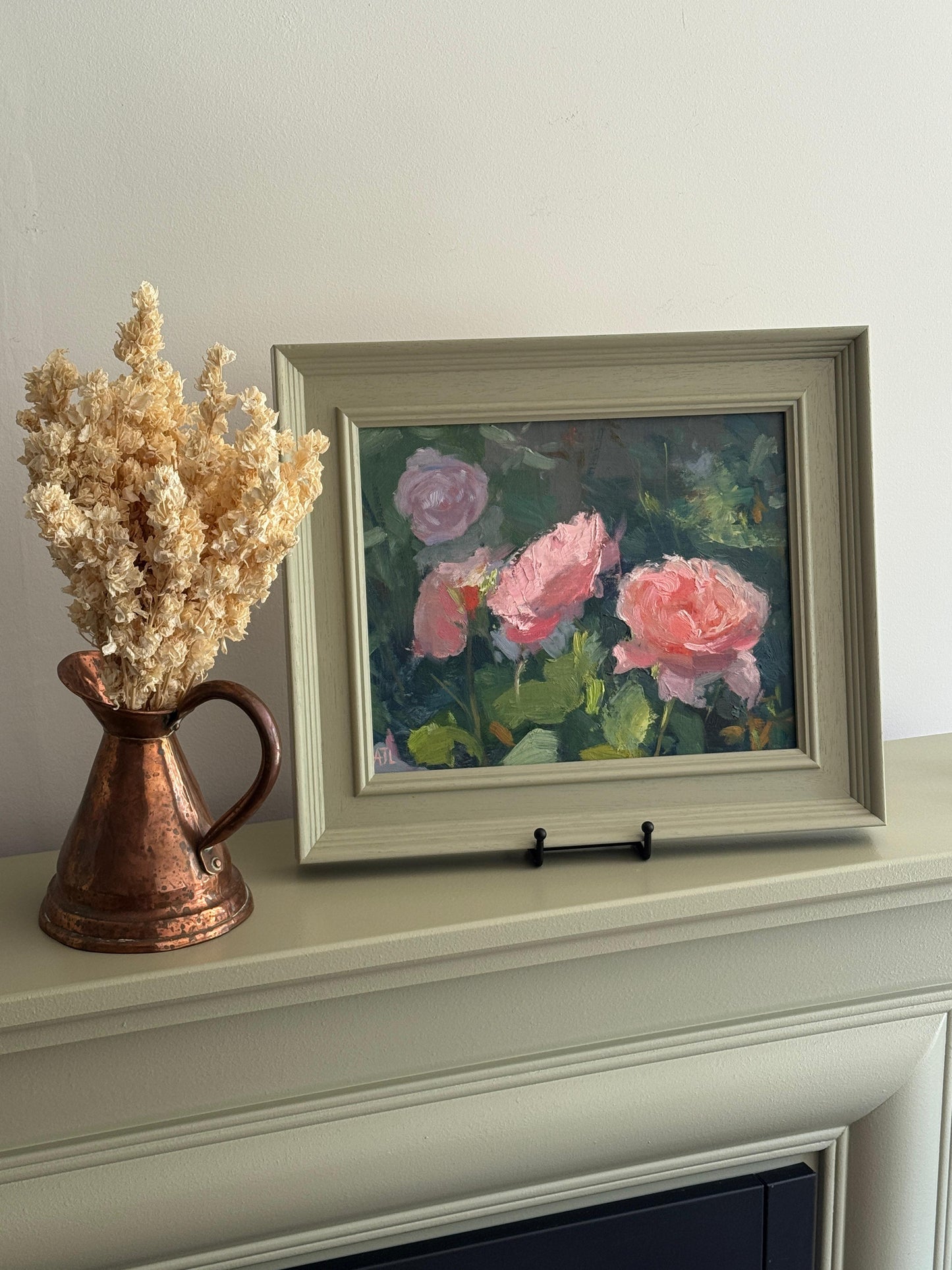 Framed painting of pink roses on a mantelpiece with a copper pitcher and dried flowers.