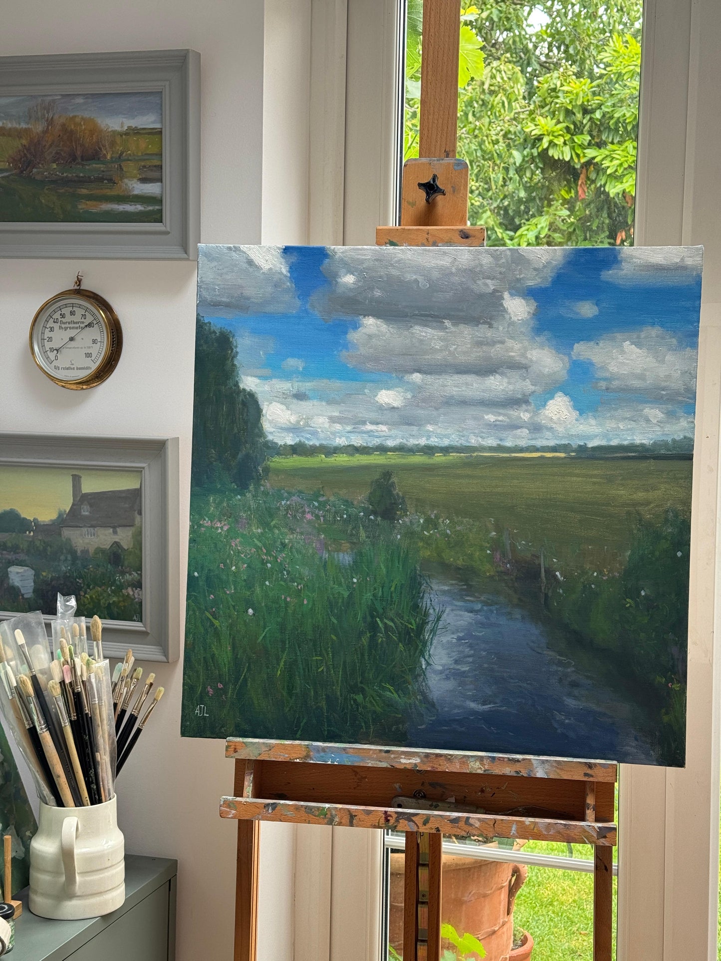 Painting of an original landscape painting on an easel in a room with framed pictures and a hygrometer on the walls.