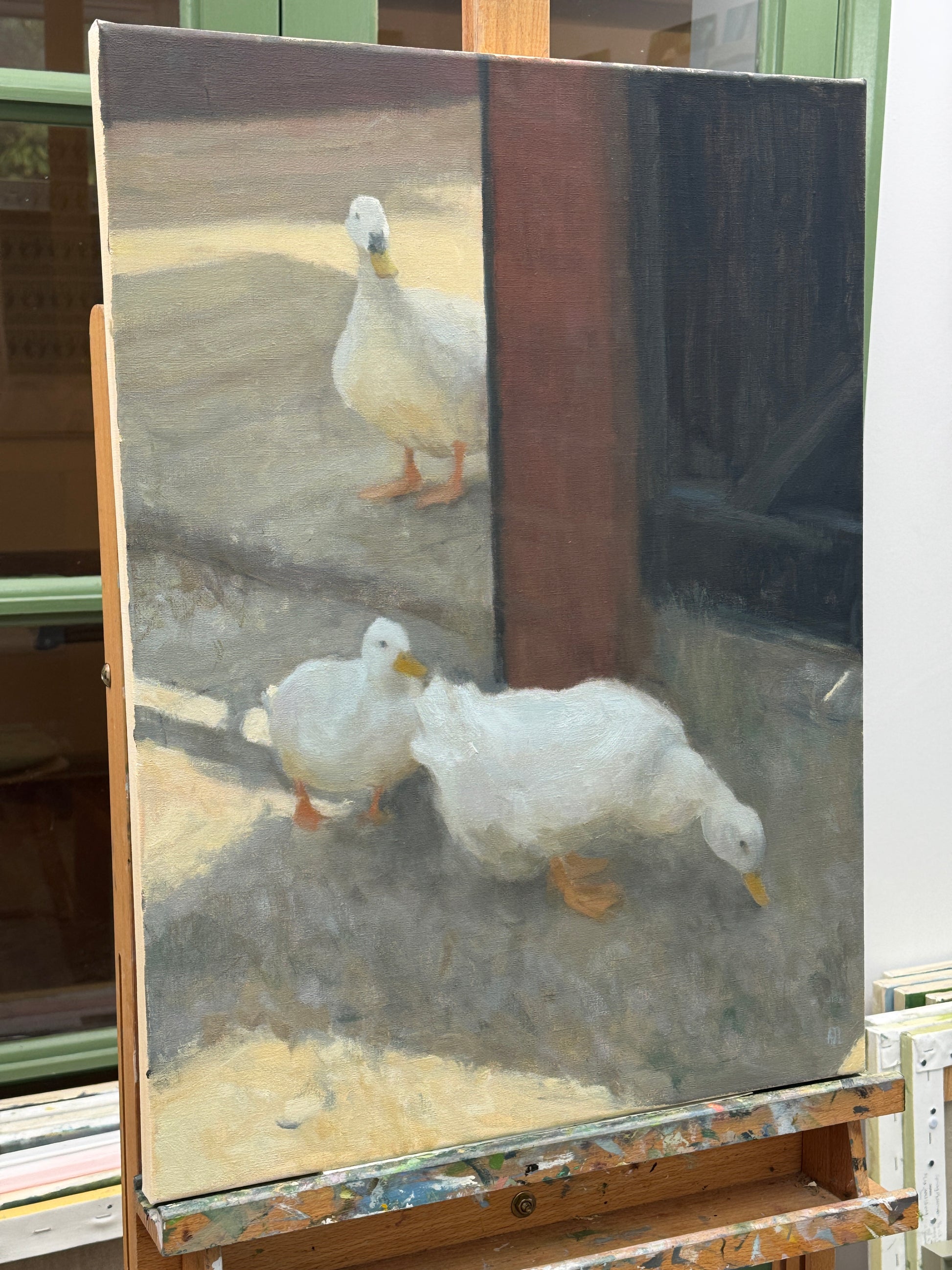 A vertical oil painting on canvas depicting three white ducks in a rustic outdoor setting, displayed on a wooden artist easel.