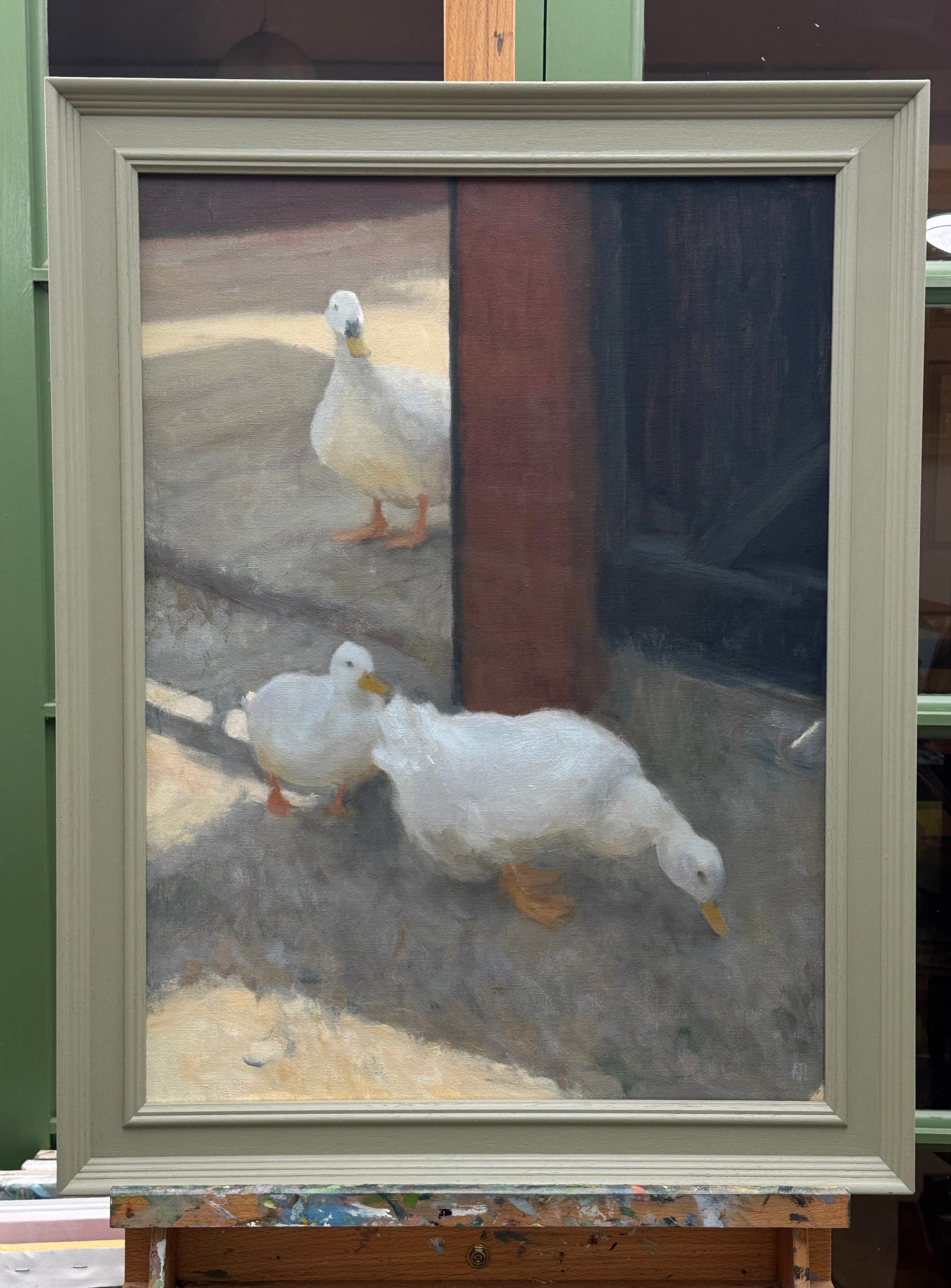 A framed oil painting of three white ducks in a courtyard, displayed on an easel. The painting features two white ducks in the foreground and a smaller one behind them near a wooden post.