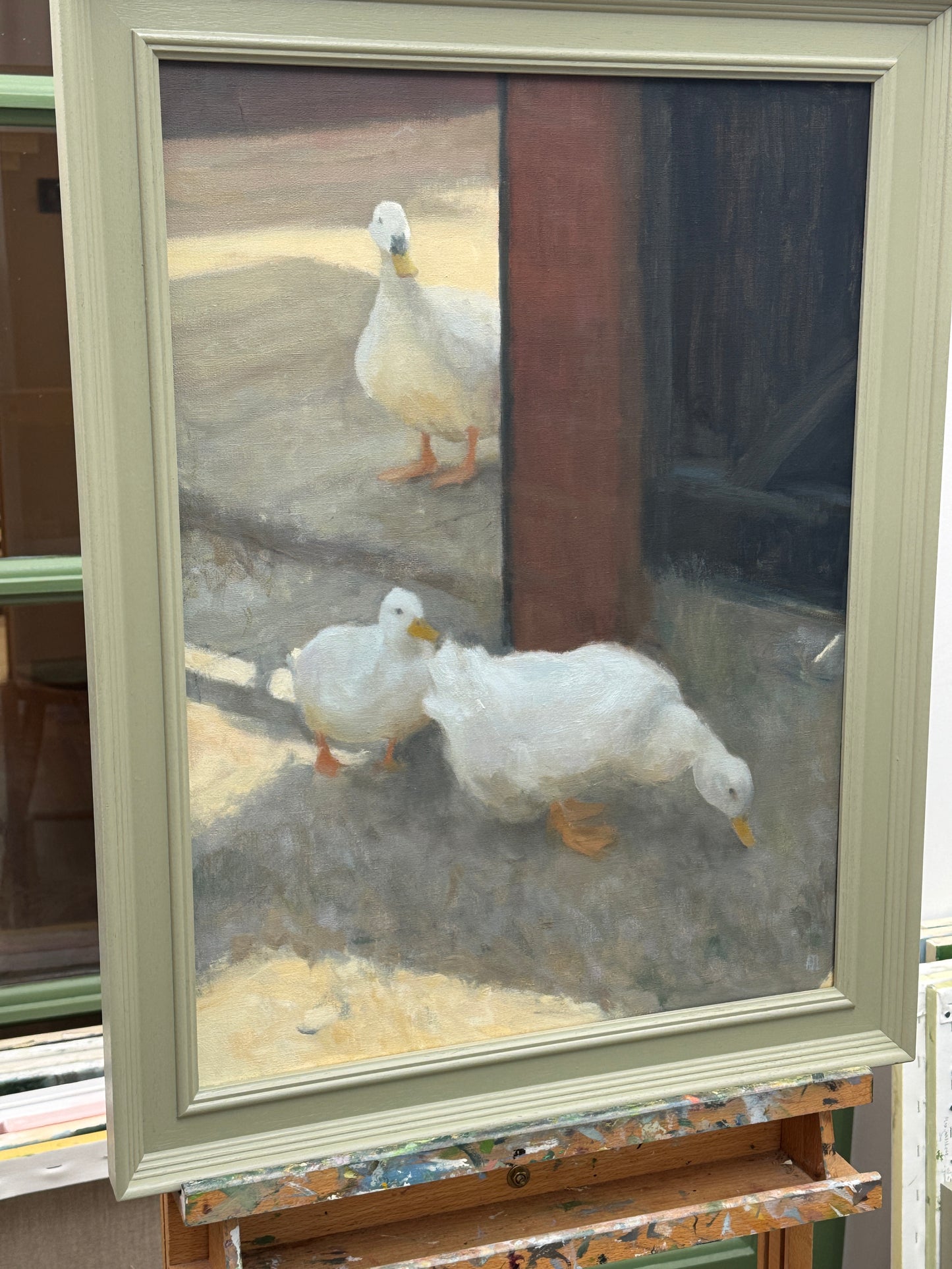 A framed oil painting depicting three white ducks in a rustic courtyard setting, displayed on an easel. The painting is housed in a light sage green wooden frame.