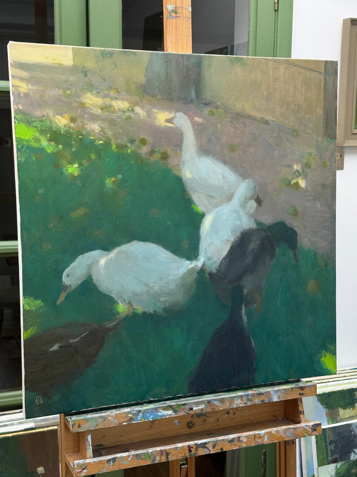 An oil painting of several ducks in a grassy outdoor setting, displayed on a wooden artist easel in an indoor studio.