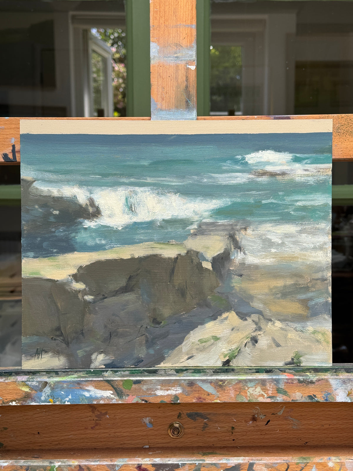 An impressionistic landscape painting of a coastal scene with teal water, white surf, and gray cliffs, shown on a wooden easel in the artist's studio.